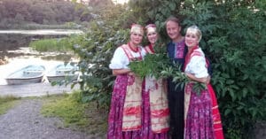 tre kvinnor och en man i ryskinspirerade dräkter framför en buske och en sjö