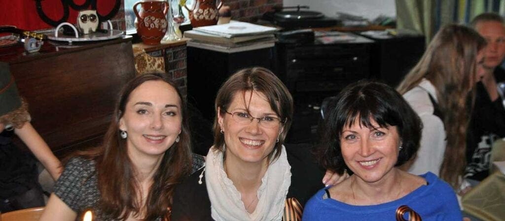 три улыбающиеся женщины, сидящие рядом друг с другом за столом и смотрящие в камеру