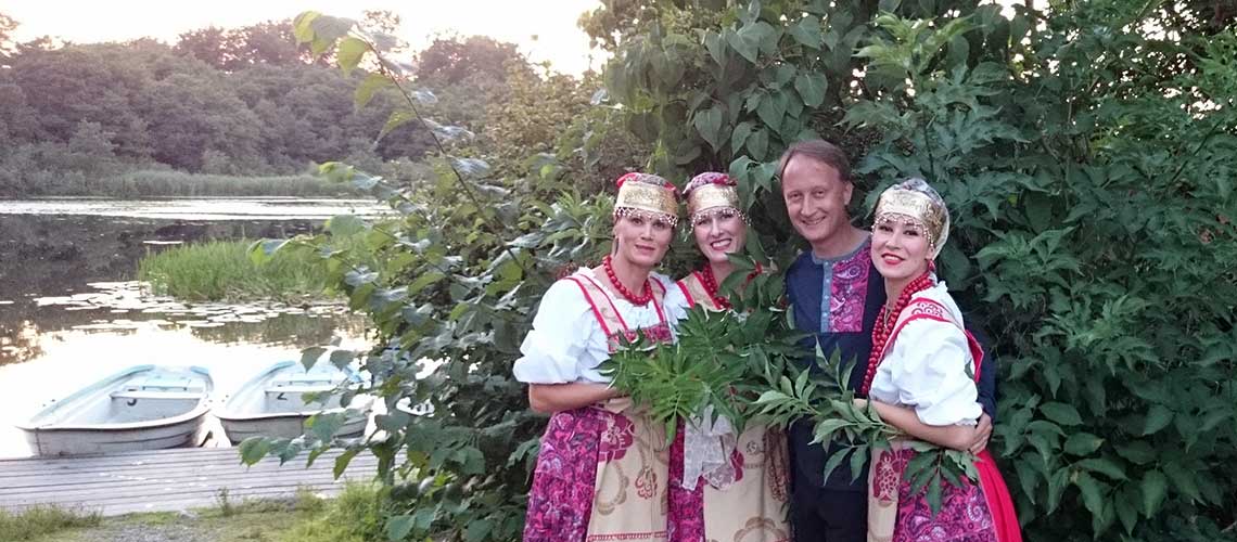 tre kvinnor och en man i ryskinspirerade dräkter framför en buske och en sjö